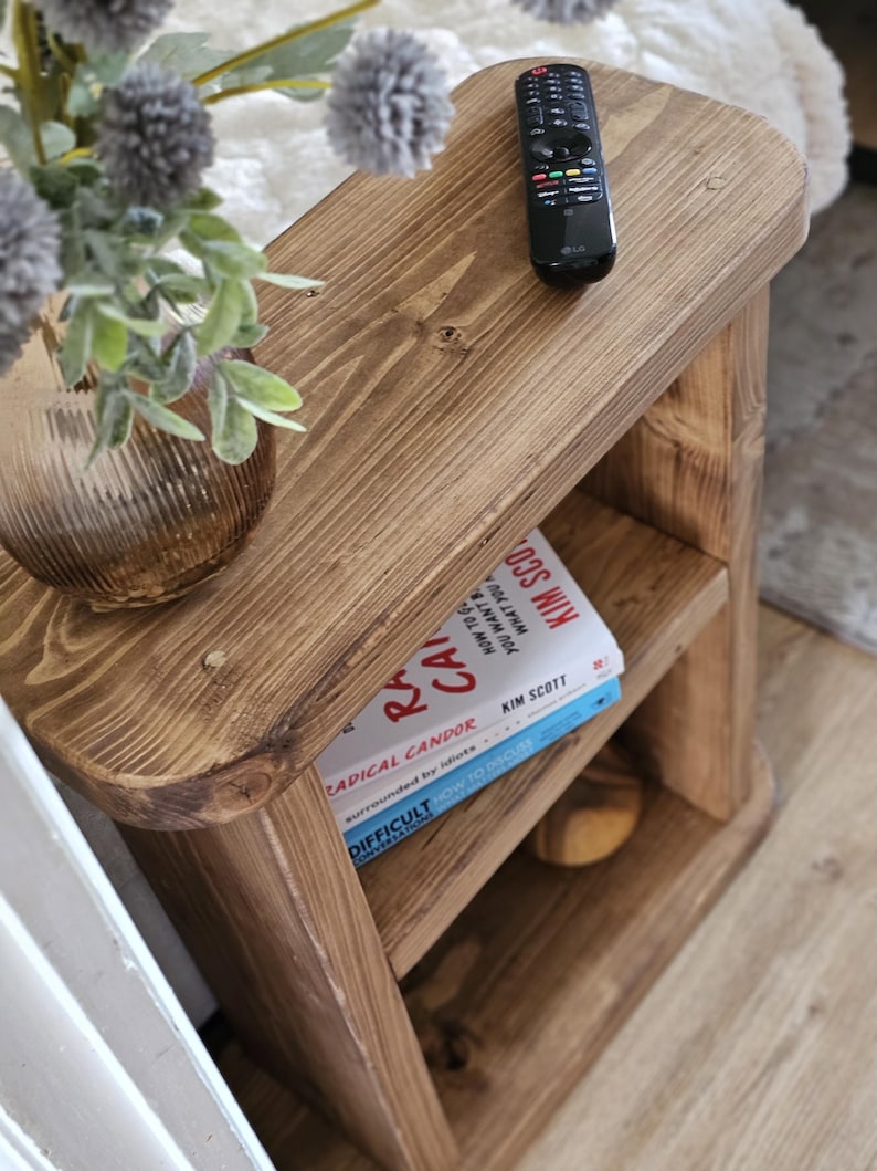 May include: A small, rustic wooden side table with two shelves. The table is stained a warm brown and has rounded edges. A black remote control sits on top, and two books are visible on the middle shelf. A vase with flowers is on the top shelf.
