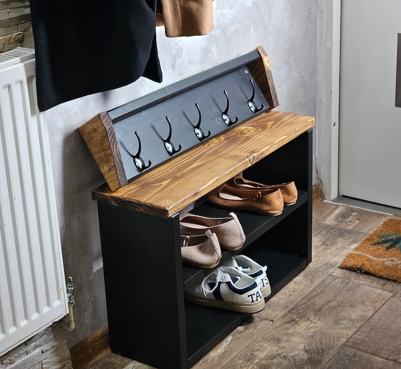 Op de afbeelding: Een zwarte houten schoenenrek met een plank en haken. Het rek heeft een bruin houten bovenblad en zwarte metalen haken. Er staan schoenen op de planken en een paar sneakers op de onderste plank.
