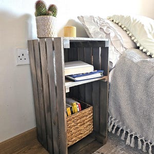 May include: A wooden crate nightstand with two shelves, a wicker basket, and books. The nightstand is painted a dark grey colour and has a rustic look. A potted cactus sits on top of the nightstand.