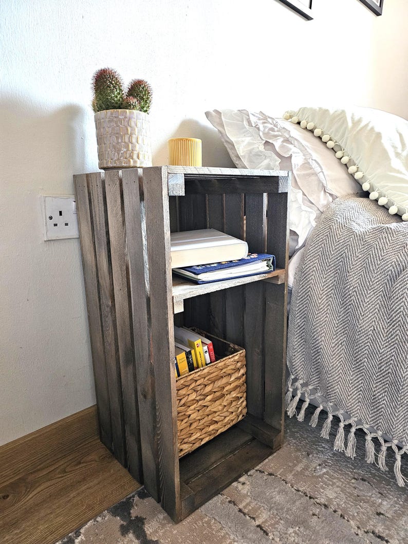 May include: A wooden crate nightstand with two shelves, a wicker basket, and books. The nightstand is painted a dark grey colour and has a rustic look. A potted cactus sits on top of the nightstand.