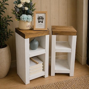 May include: Two white wooden side tables with a dark brown top. One table has a vase, books, and a framed picture of a cat wearing glasses. The other table is empty. The tables are on a light brown wooden floor.