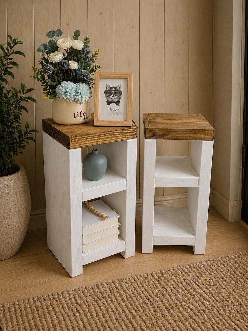 May include: Two white wooden side tables with a dark brown top. One table has a vase, books, and a framed picture of a cat wearing glasses. The other table is empty. The tables are on a light brown wooden floor.