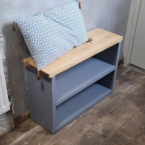 May include: A gray wooden shoe rack with two shelves and a natural wood top. A blue and white patterned pillow with tassels sits on top of the rack.