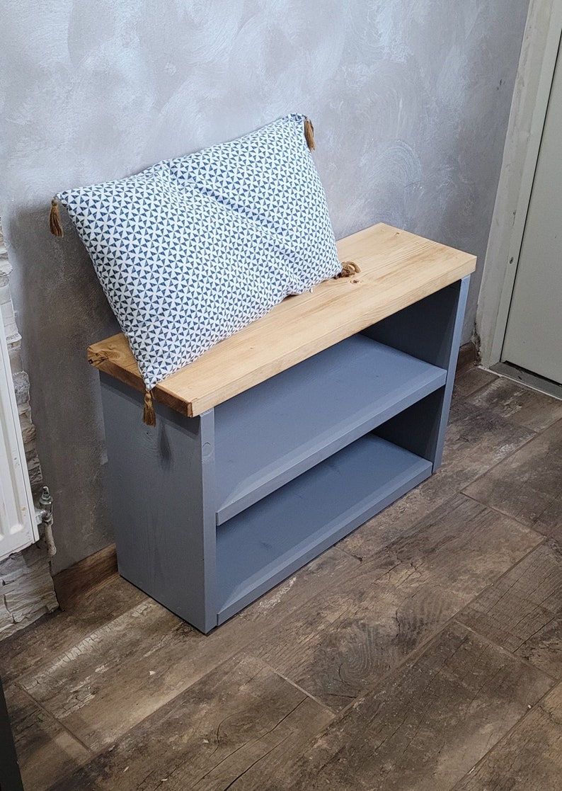 May include: A gray wooden shoe rack with two shelves and a natural wood top. A blue and white patterned pillow with tassels sits on top of the rack.