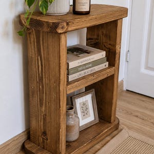 May include: A rustic, wooden side table with two shelves. The top shelf holds a potted plant and a scented candle. The middle shelf displays books, and the bottom shelf features a framed picture and a small vase. The table is made of stained wood.