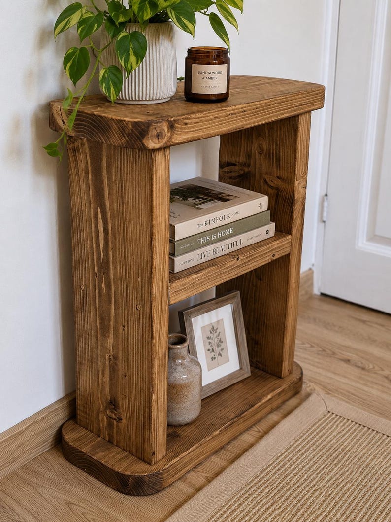 May include: A rustic, wooden side table with two shelves. The top shelf holds a potted plant and a scented candle. The middle shelf displays books, and the bottom shelf features a framed picture and a small vase. The table is made of stained wood.