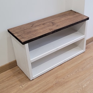 May include: White wooden shoe rack with two shelves and a brown stained wood top. The shoe rack is sitting on a wood floor.