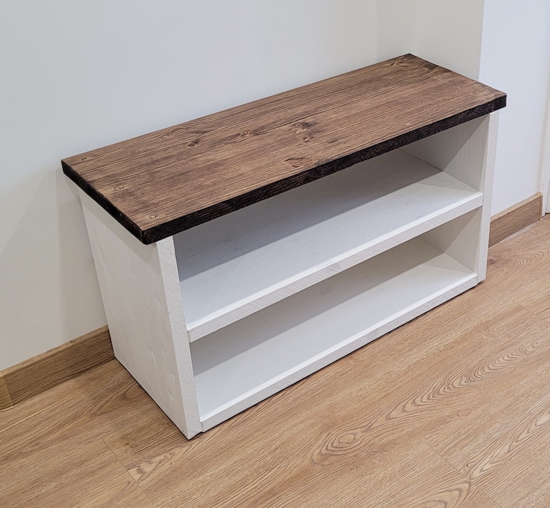May include: White wooden shoe rack with two shelves and a brown stained wood top. The shoe rack is sitting on a wood floor.