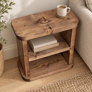 May include: A rectangular wooden side table with a dark brown finish and visible wood grain. The table features a flat top surface, a middle shelf holding two stacked books, and a solid base. A ceramic mug sits on a coaster on the top surface.