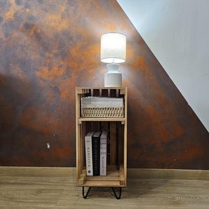 May include: A wooden crate nightstand with two shelves. The top shelf has a white wicker basket and a white lamp with a white shade. The bottom shelf has three books: Kinfolk, The Style of Giving, and Elle. The nightstand has black hairpin legs and is sitting on a wood floor.