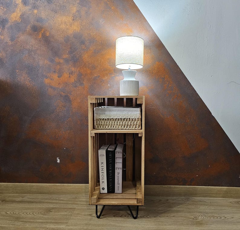 May include: A wooden crate nightstand with two shelves. The top shelf has a white wicker basket and a white lamp with a white shade. The bottom shelf has three books: Kinfolk, The Style of Giving, and Elle. The nightstand has black hairpin legs and is sitting on a wood floor.