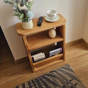 May include: A wooden shelf unit with three shelves. The top shelf has a white ceramic cup and saucer, the middle shelf has a ceramic vase with flowers, and the bottom shelf has books and a magazine. The shelf is on a wooden floor with a patterned rug.