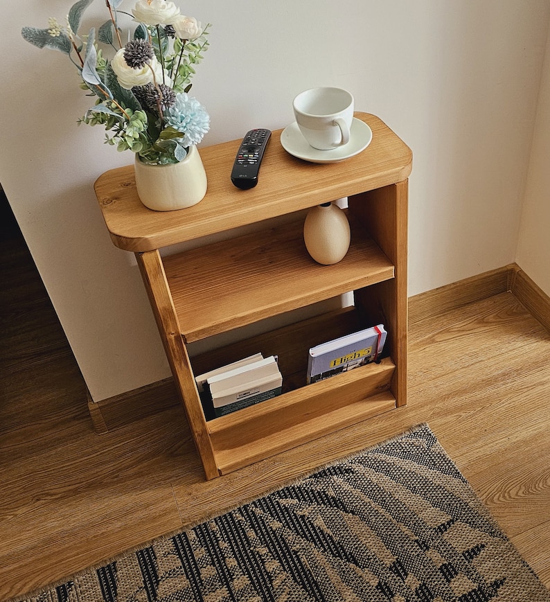 May include: A wooden shelf unit with three shelves. The top shelf has a white ceramic cup and saucer, the middle shelf has a ceramic vase with flowers, and the bottom shelf has books and a magazine. The shelf is on a wooden floor with a patterned rug.