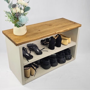 May include: A white wooden shoe rack with two shelves. The top shelf is made of brown wood and has a plant on it. The bottom shelf is filled with shoes, including sneakers, sandals, and heels.