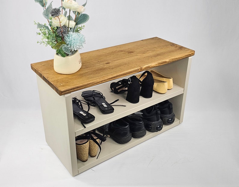 May include: A white wooden shoe rack with two shelves. The top shelf is made of brown wood and has a plant on it. The bottom shelf is filled with shoes, including sneakers, sandals, and heels.