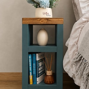 May include: A teal bedside table with a wooden top, holding a vase of flowers and books. The vase has the word "Life" on it. The table has two shelves and sits on a patterned rug. A bed with white linens is in the background.