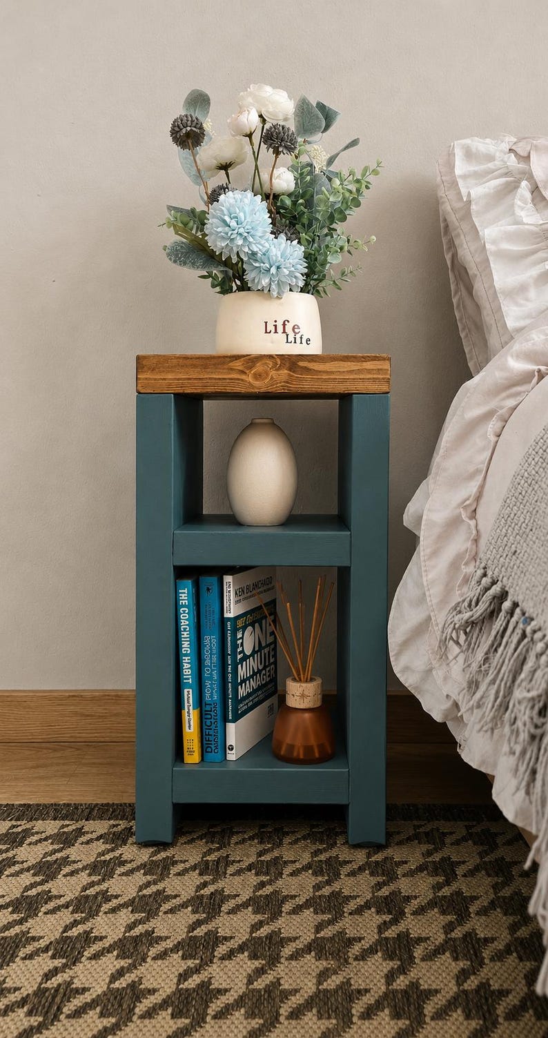 May include: A teal bedside table with a wooden top, holding a vase of flowers and books. The vase has the word "Life" on it. The table has two shelves and sits on a patterned rug. A bed with white linens is in the background.
