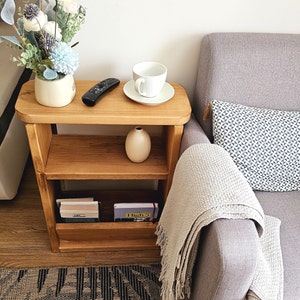 May include: A wooden side table with two shelves, a white ceramic vase, a white coffee cup and saucer, a black remote control, and a stack of books. The table is next to a gray sofa with a white throw blanket and a patterned pillow.