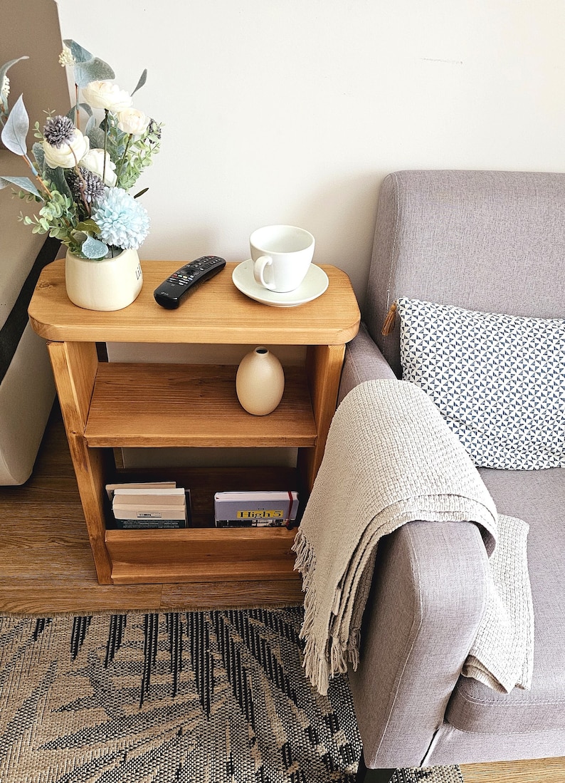 May include: A wooden side table with two shelves, a white ceramic vase, a white coffee cup and saucer, a black remote control, and a stack of books. The table is next to a gray sofa with a white throw blanket and a patterned pillow.