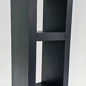 May include: A tall, black wooden shelf with two interior shelves and a natural wood top. The shelf is rectangular with a modern design, perfect for displaying items or storage. The wood grain is visible on the top shelf.