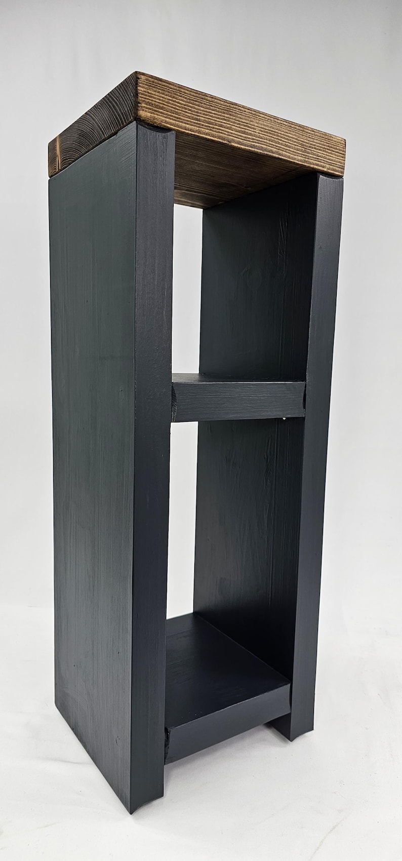 May include: A tall, black wooden shelf with two interior shelves and a natural wood top. The shelf is rectangular with a modern design, perfect for displaying items or storage. The wood grain is visible on the top shelf.