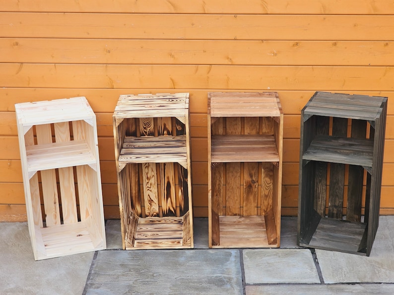 May include: Four wooden crates with shelves, each finished in a different colour: natural wood, burnt wood, stained wood, and dark grey. The crates are arranged in a row on a stone floor in front of a wooden wall.
