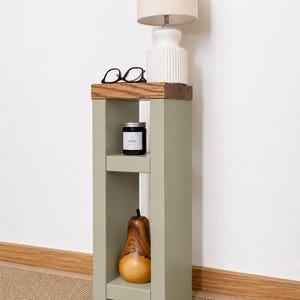 May include: A sage green side table with a wooden top, holding a white lamp with a beige lampshade, black glasses, and a black candle labeled "calm." A wooden pear decorates the lower shelf.
