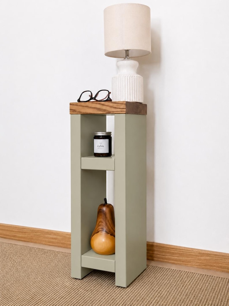 May include: A sage green side table with a wooden top, holding a white lamp with a beige lampshade, black glasses, and a black candle labeled "calm." A wooden pear decorates the lower shelf.