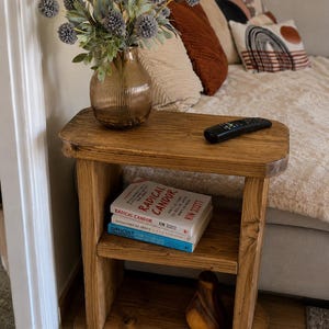 May include: A rustic wooden side table with two shelves. A vase of gray flowers sits on top, along with a remote control. The bottom shelf holds two books and a decorative wooden pear. The table is stained a warm brown.