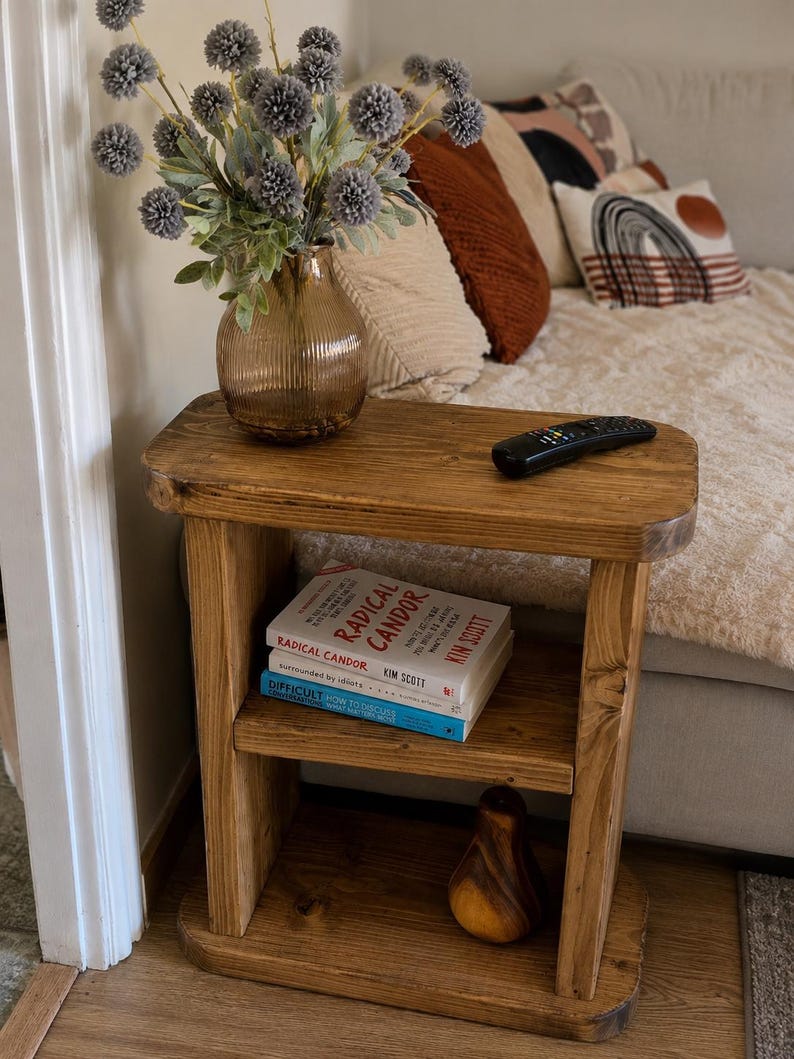 May include: A rustic wooden side table with two shelves. A vase of gray flowers sits on top, along with a remote control. The bottom shelf holds two books and a decorative wooden pear. The table is stained a warm brown.