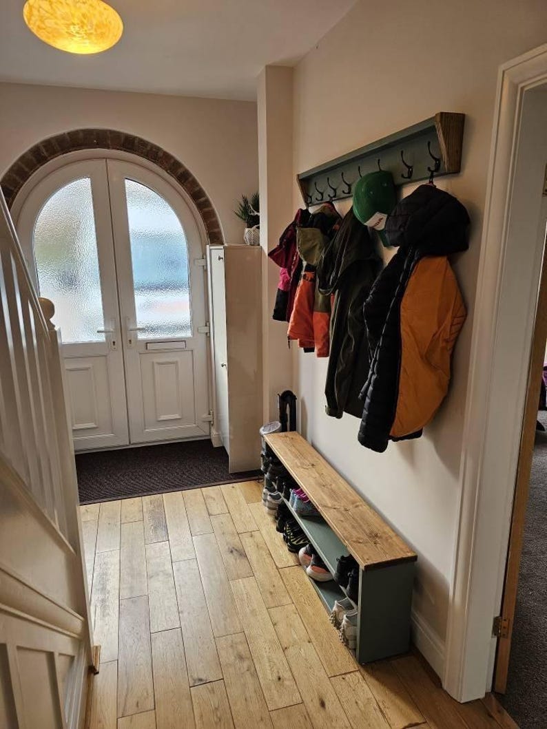May include: A green wooden coat rack with hooks and a wooden shelf underneath. The rack is mounted on a white wall and has several coats and hats hanging on it. There is a wooden bench with a shelf underneath, and the bench is filled with shoes.