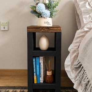 May include: A black and brown wooden side table with a shelf, holding books, a vase, and a diffuser. The top shelf displays a vase with blue and white flowers. The vase has the word "Life" on it.