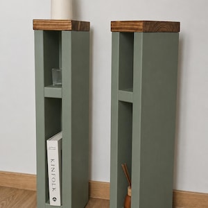 May include: Two sage green wooden side tables with a dark brown top. Each table has a rectangular shape with a shelf. One table holds a book, the other a small bottle with reeds. The tables are on a wooden floor.