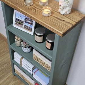 A wooden painted bookshelf with candles, framed photos, two baskets, decorative jars, and stacked books  placed on a wooden floor against a white wall.
