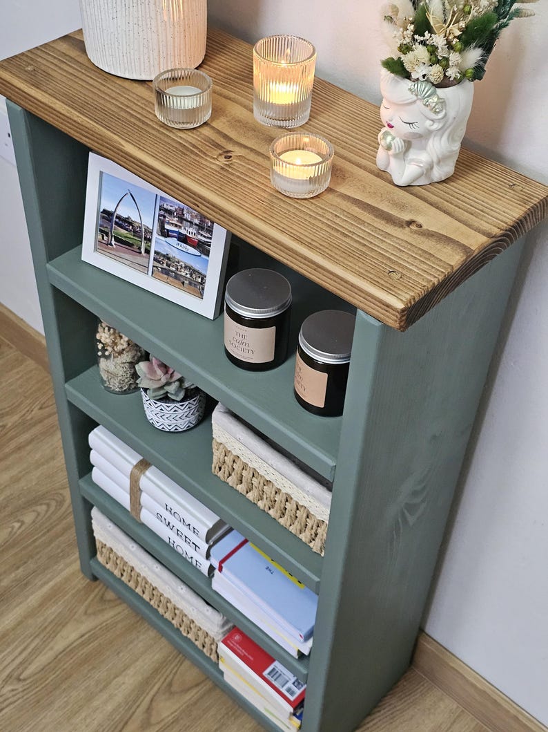 A wooden painted bookshelf with candles, framed photos, two baskets, decorative jars, and stacked books  placed on a wooden floor against a white wall.