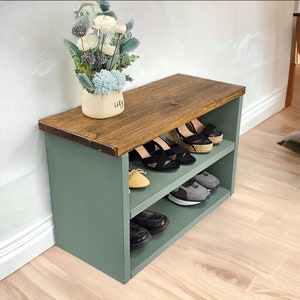 May include: A two-tiered shoe rack with a wooden top and green painted sides. The top shelf is made of wood and the bottom shelf is made of metal. The rack is filled with shoes, including sandals, sneakers, and loafers.