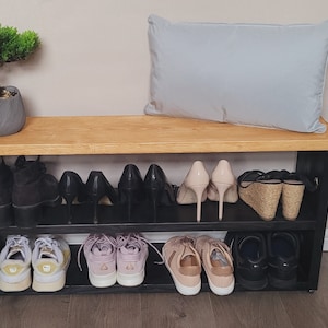 May include: A black shoe rack with two shelves, a wooden top, and a light gray pillow on top. The shoe rack is filled with various shoes, including sneakers, heels, and sandals.
