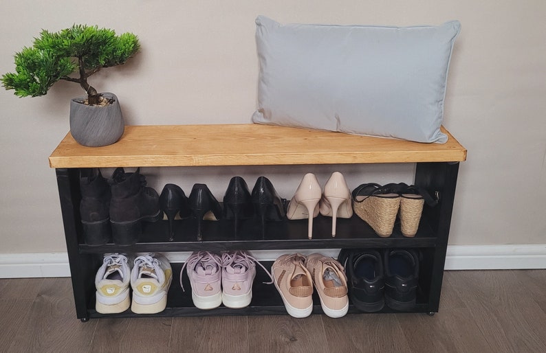 May include: A black shoe rack with two shelves, a wooden top, and a light gray pillow on top. The shoe rack is filled with various shoes, including sneakers, heels, and sandals.
