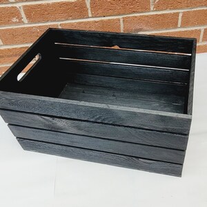 Puede incluir: Una caja de madera negra con un asa en el lateral. La caja tiene cuatro listones en la parte delantera y los laterales.