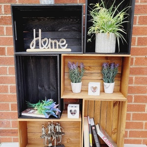 Puede incluir: Una estanter&iacute;a de madera de color natural y negro con tres estantes. El estante superior tiene una peque&ntilde;a planta en maceta y la palabra "Home" en letras decorativas. El estante del medio tiene dos peque&ntilde;as plantas en maceta y un portavelas blanco en forma de coraz&oacute;n. El estante inferior tiene una peque&ntilde;a escultura de personas sentadas en un banco, un libro y una pila de libros con la palabra "Kaizen" en el libro de arriba.