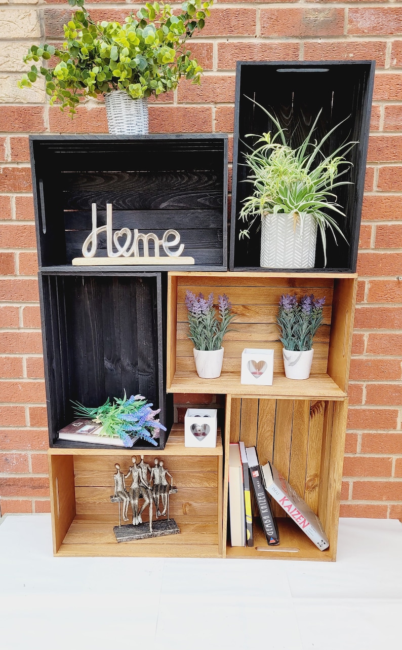 Puede incluir: Una estanter&iacute;a de madera de color natural y negro con tres estantes. El estante superior tiene una peque&ntilde;a planta en maceta y la palabra "Home" en letras decorativas. El estante del medio tiene dos peque&ntilde;as plantas en maceta y un portavelas blanco en forma de coraz&oacute;n. El estante inferior tiene una peque&ntilde;a escultura de personas sentadas en un banco, un libro y una pila de libros con la palabra "Kaizen" en el libro de arriba.