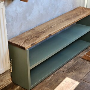 Op de afbeelding: Een groene houten schoenenrek met twee planken en een houten bovenblad. Het bovenblad is donkerbruin gebeitst. Het schoenenrek staat op een betegelde vloer.