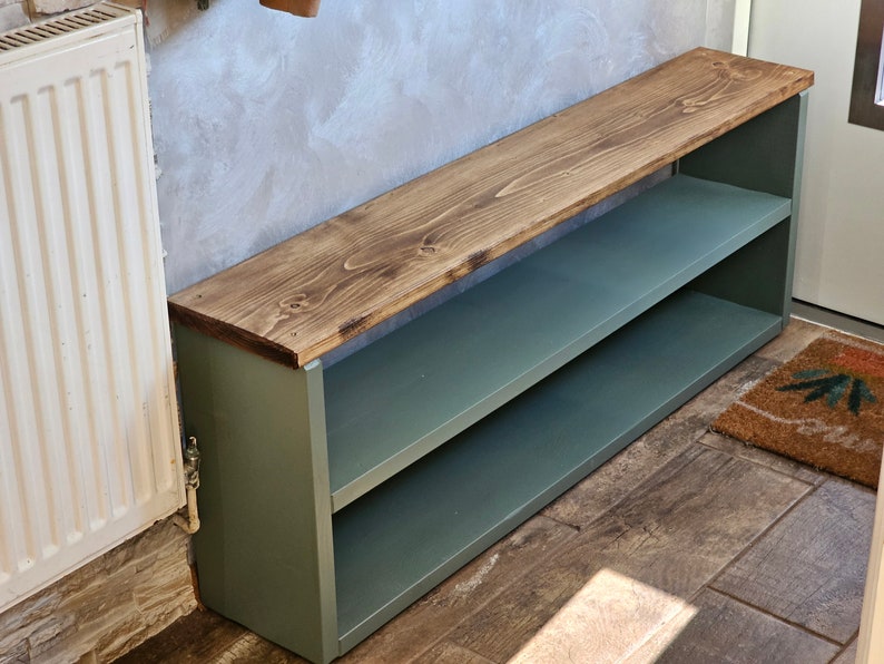 Op de afbeelding: Een groene houten schoenenrek met twee planken en een houten bovenblad. Het bovenblad is donkerbruin gebeitst. Het schoenenrek staat op een betegelde vloer.