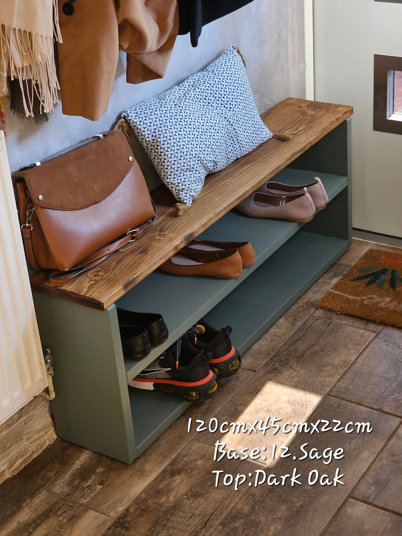 May include: A wooden shoe bench with two shelves, painted sage green with a dark oak top. The bench is 47 inches long, 18 inches wide, and 9 inches tall. It has a cushion on top and is decorated with a brown leather bag, a patterned pillow, and shoes.