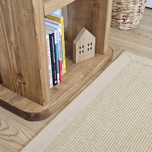 May include: A wooden bookshelf with books, a small wooden house, and a beige rug. The bookshelf is made of light brown wood and has several books on the shelf. The rug is rectangular with a beige border.