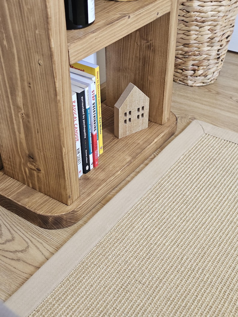 May include: A wooden bookshelf with books, a small wooden house, and a beige rug. The bookshelf is made of light brown wood and has several books on the shelf. The rug is rectangular with a beige border.