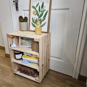 May include: A wooden shelf unit with four shelves, featuring a framed print of a lemon tree with the word "CITRON" on it, a small potted cactus, a candle, a book, and a decorative box. The shelf unit is on wheels and has a natural wood finish.