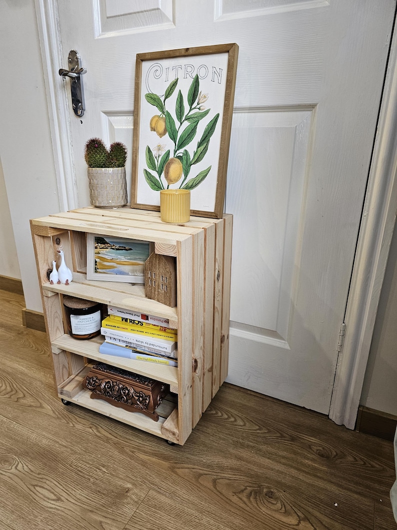 May include: A wooden shelf unit with four shelves, featuring a framed print of a lemon tree with the word "CITRON" on it, a small potted cactus, a candle, a book, and a decorative box. The shelf unit is on wheels and has a natural wood finish.