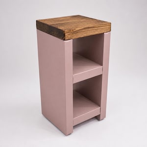 May include: A small, rectangular side table with a light brown, wooden top and a dusty rose-colored base. The base features two open shelves. The table is approximately 24 inches tall.