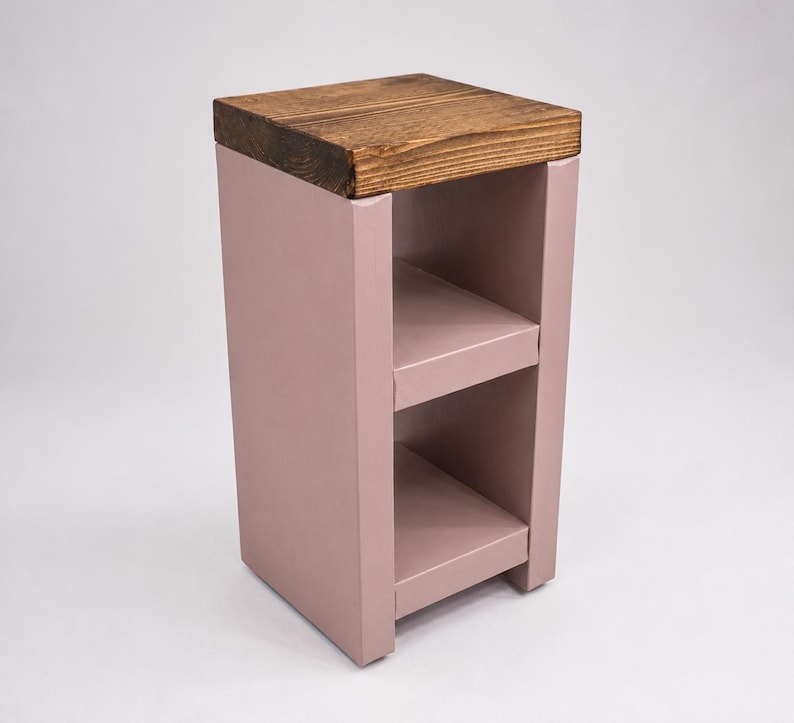 May include: A small, rectangular side table with a light brown, wooden top and a dusty rose-colored base. The base features two open shelves. The table is approximately 24 inches tall.